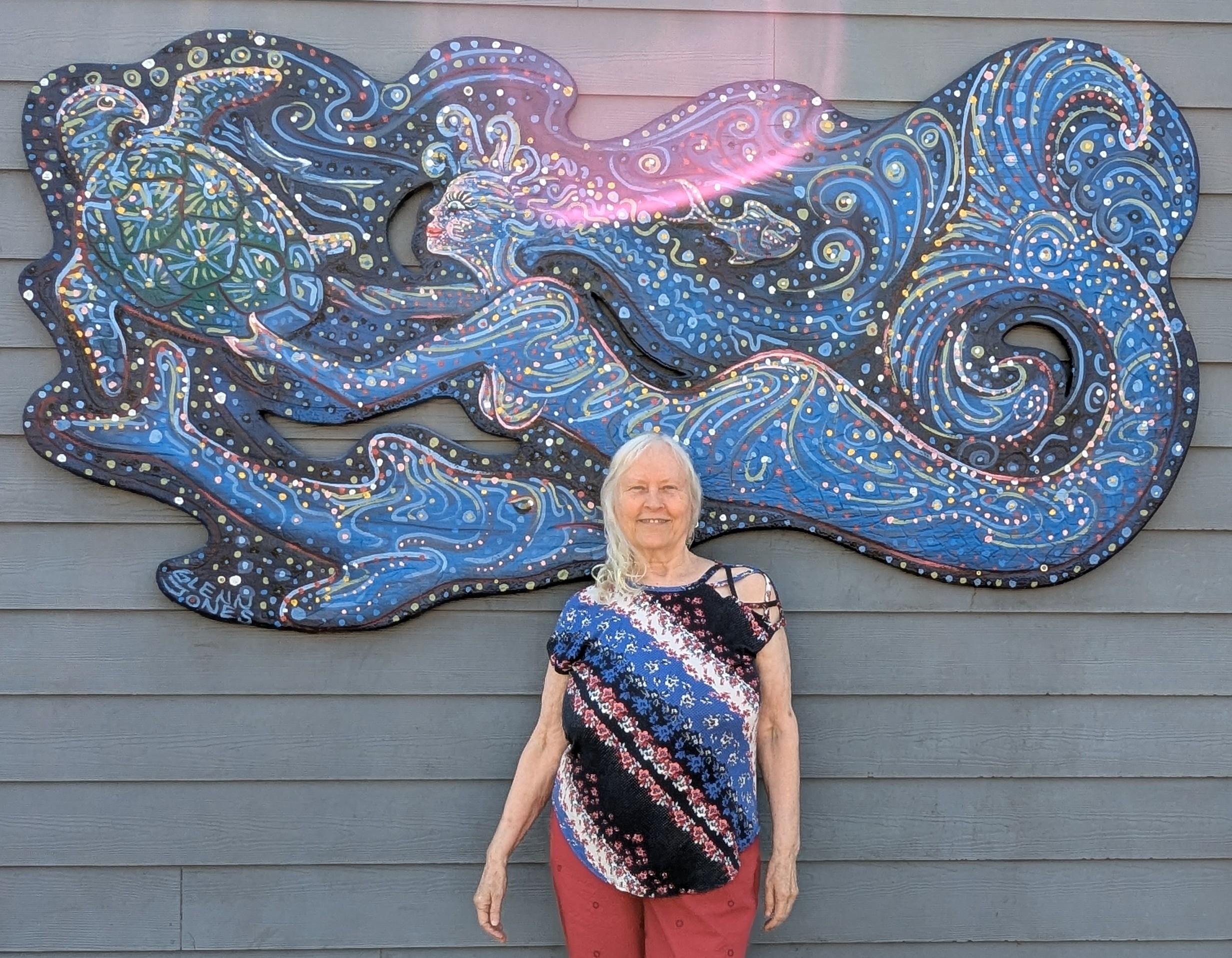 Dee Wirak in front of a woodan sign of a mermaid, a dolphin, and a turtle.